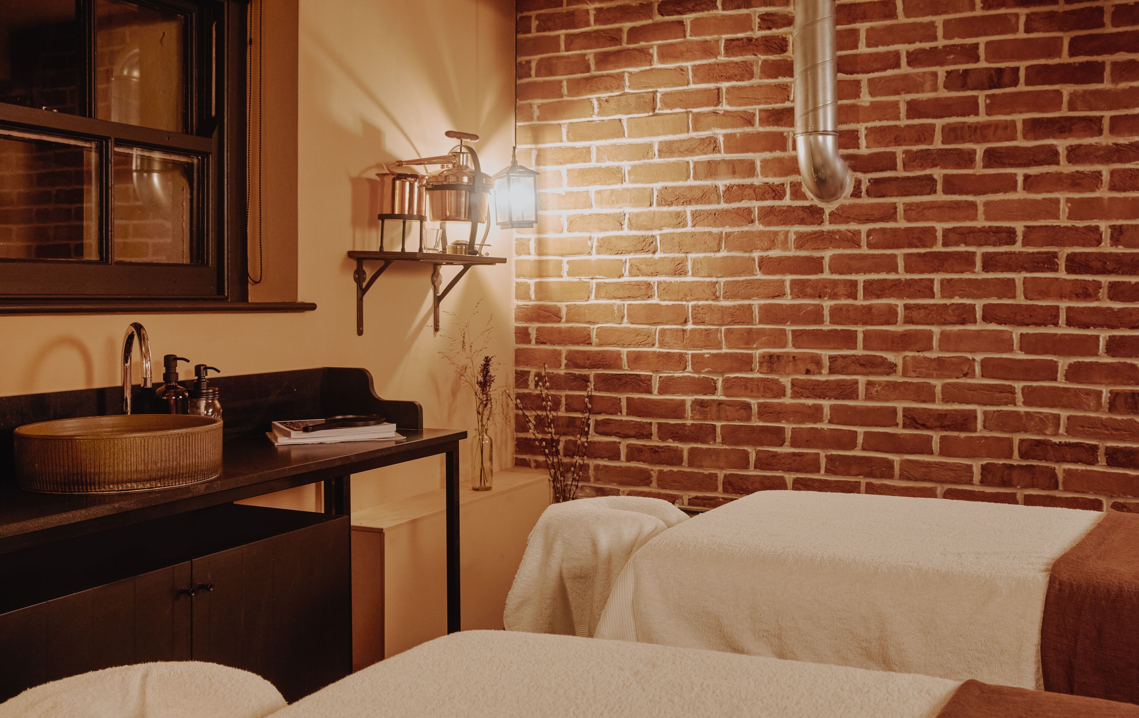 A bathroom with a brick wall, a wooden bench, a sink with toiletries, and towels on the floor.