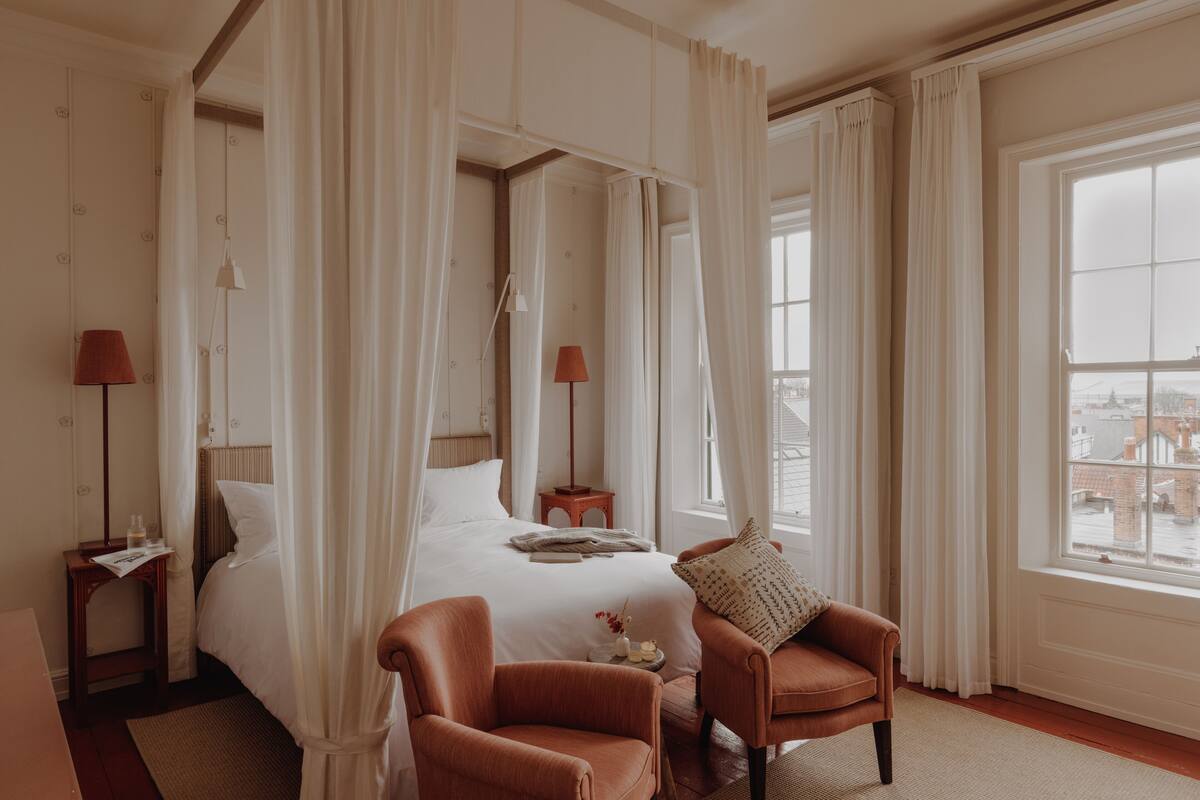A bedroom with a four-poster bed, a sitting area with a chair, and a view of the city through a large window.