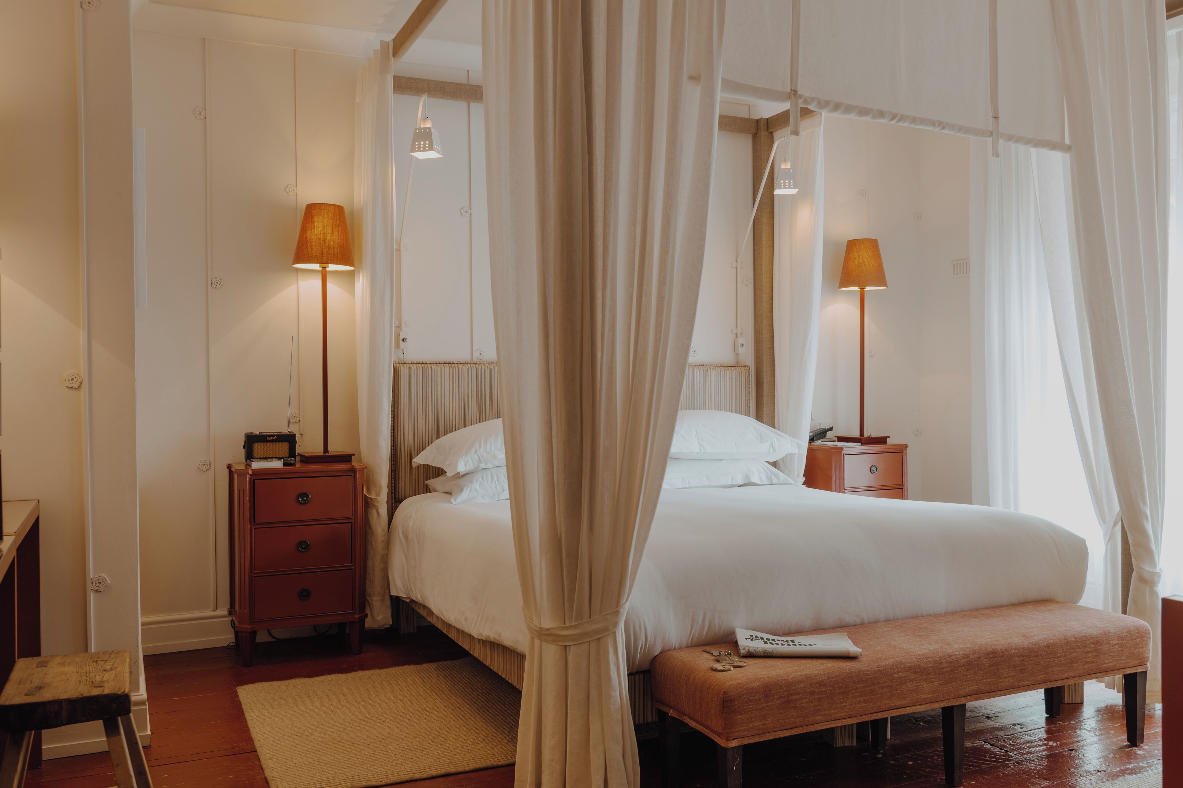 Een slaapkamer met een hemelbed, bedlampen, een houten nachtkastje en een bankje.