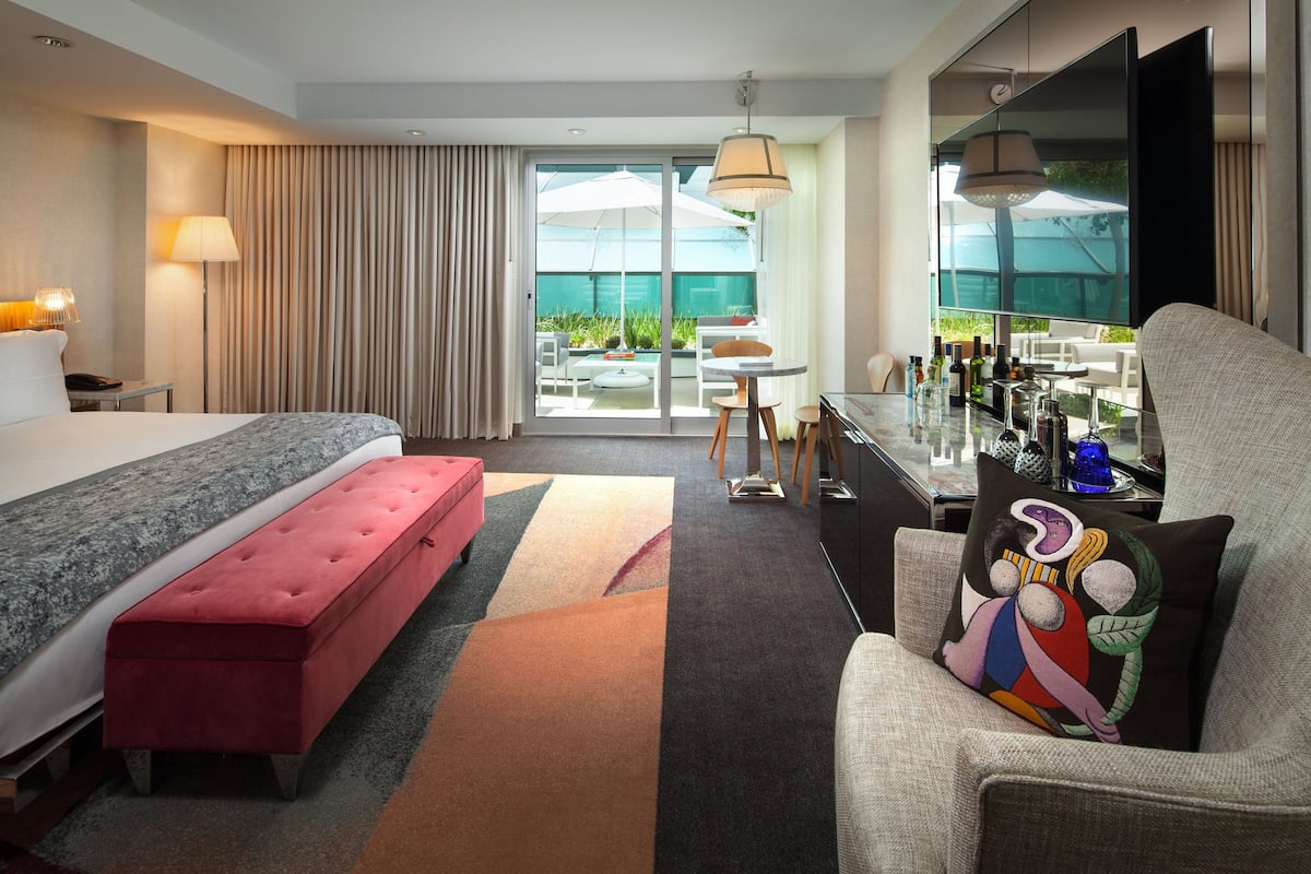 A hotel room with a bed, a red bench, a dining table, and a TV.