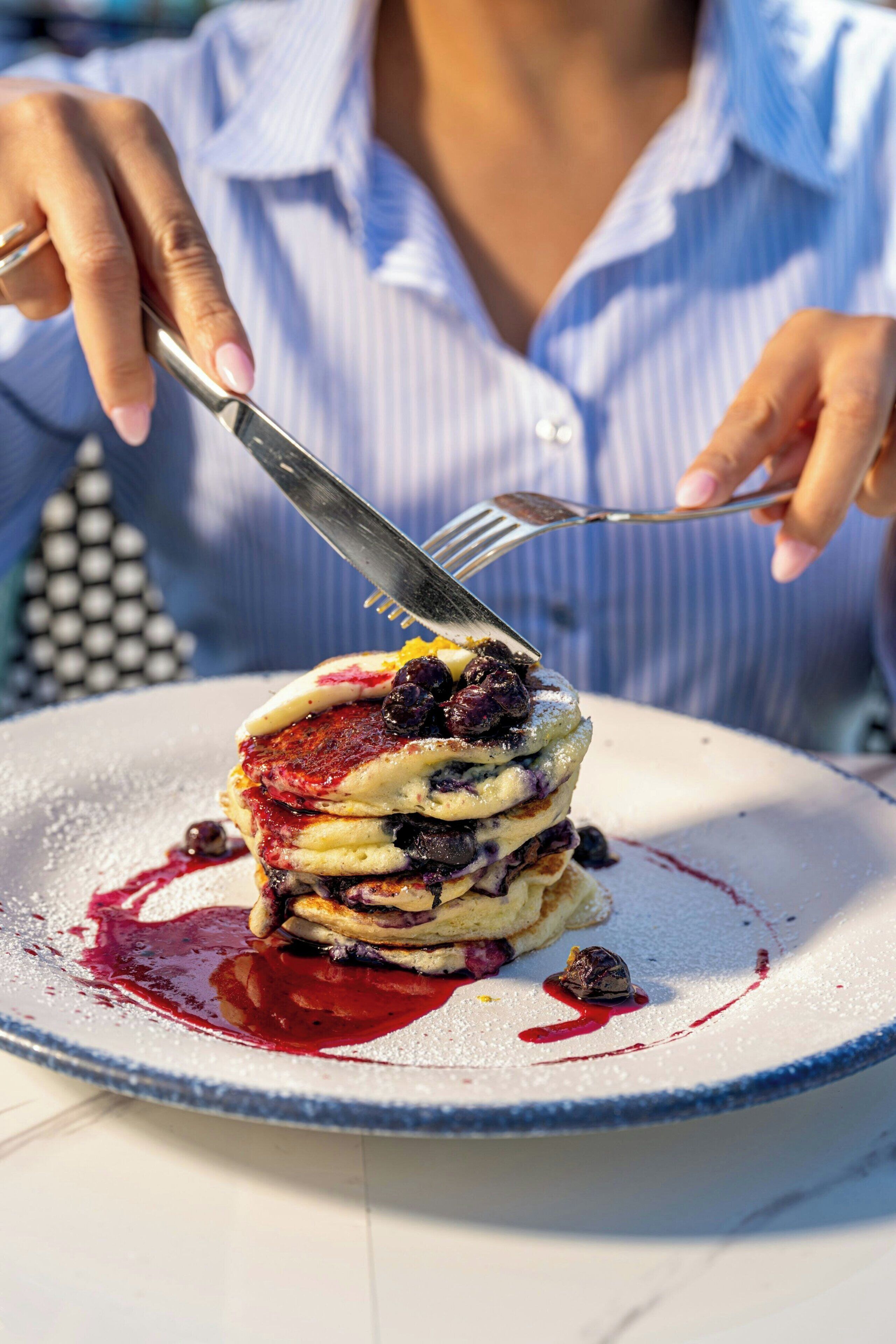 Une personne qui découpe une pile de crêpes garnies de myrtilles sur une assiette.
