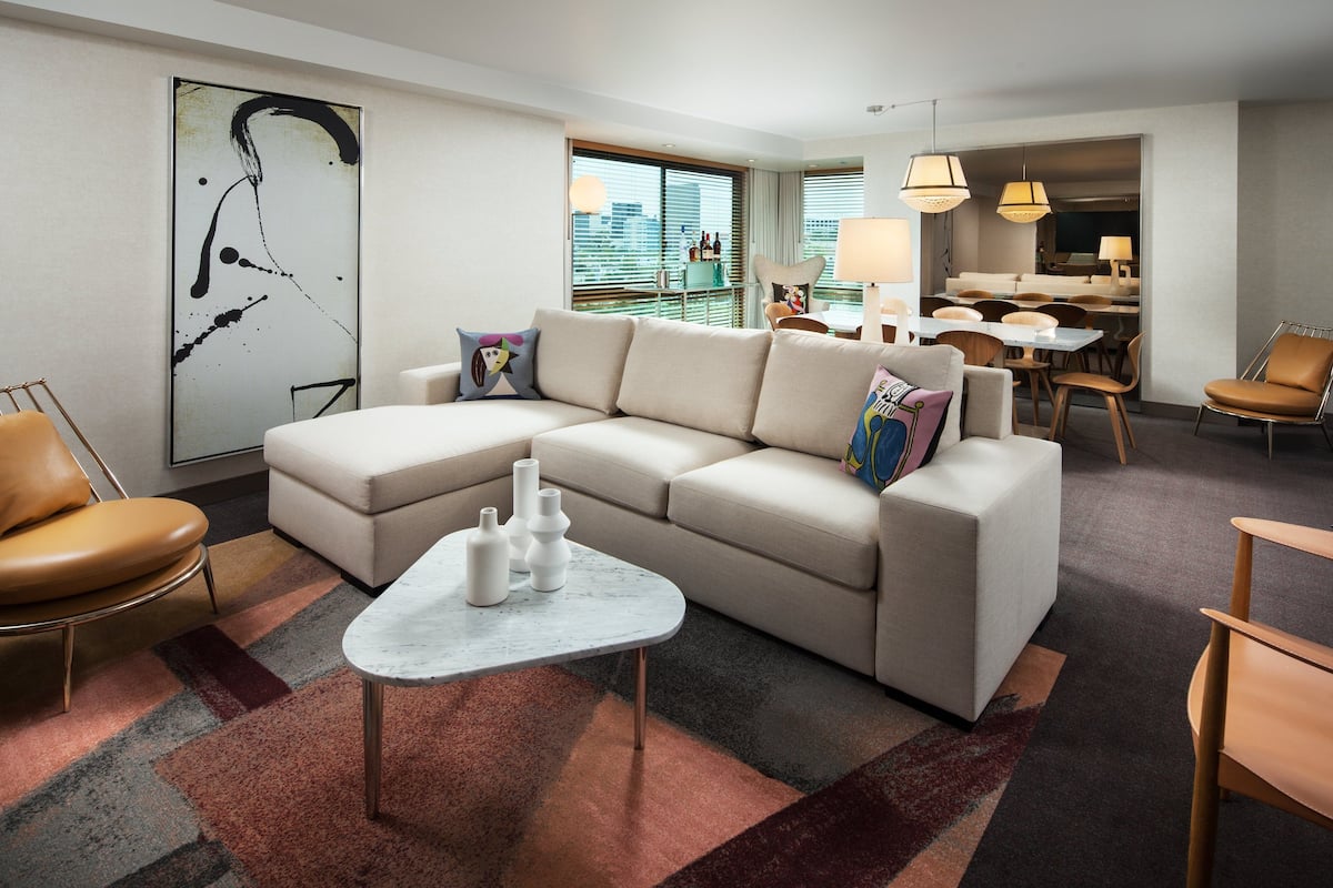 A modern hotel room with a sectional sofa, a small coffee table, and a dining area with a table and chairs.