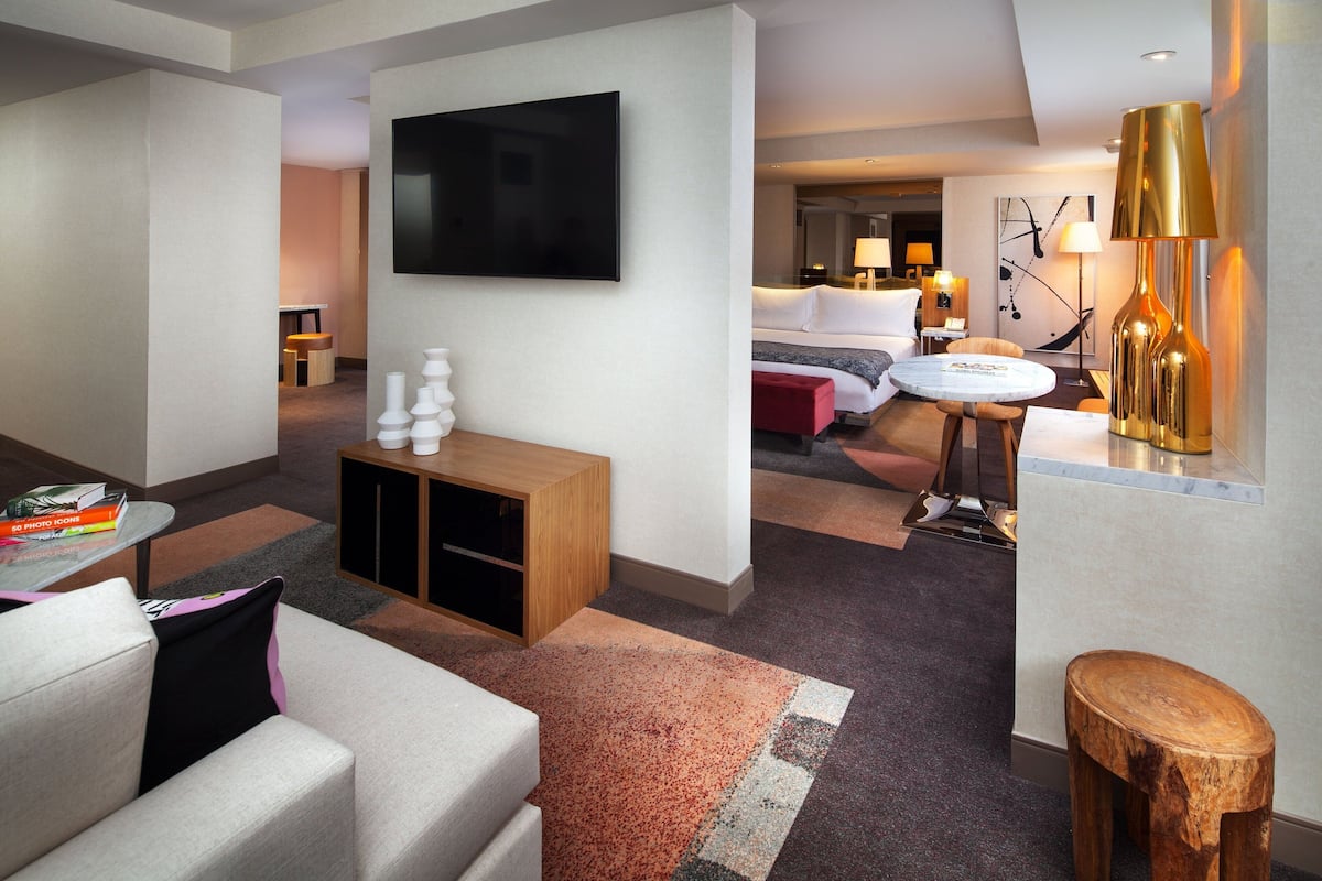 A modern hotel room with a flat-screen TV, a wooden cabinet, a round table, and a red ottoman.