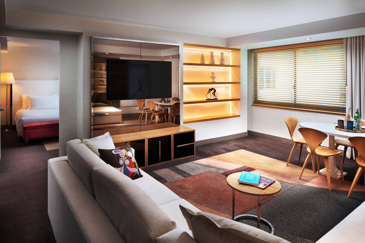 Ein modernes Hotelzimmer mit Wohnbereich, Esstisch, Sofa, Fernseher und Bücherregal.