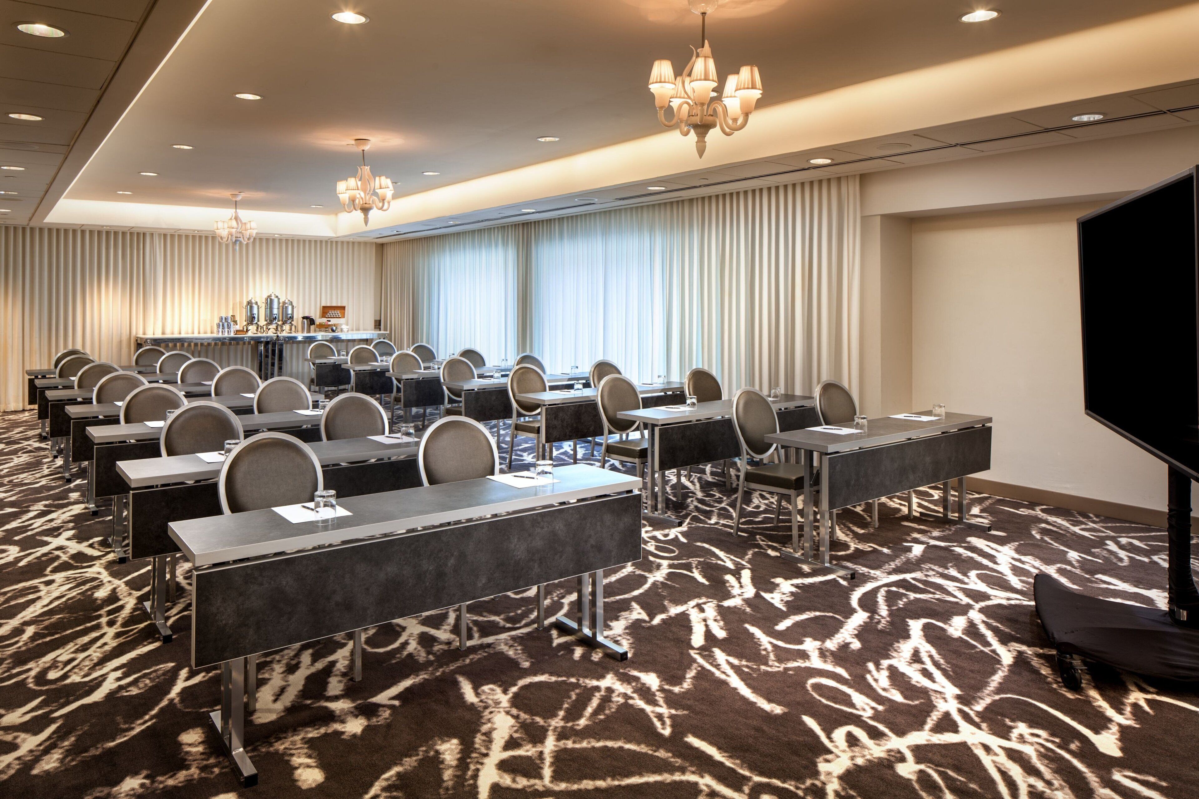 A conference room with rows of tables and chairs, a large screen, and a bar area with bottles.