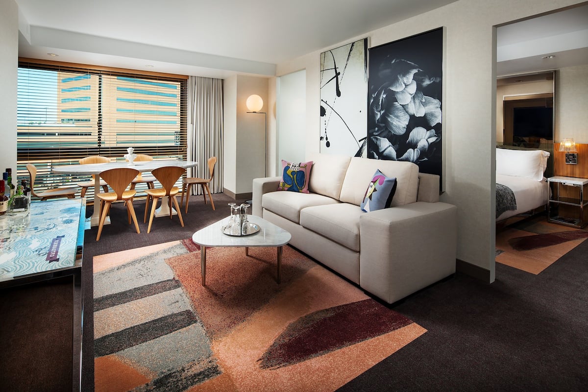 A modern hotel room with a sofa, a dining table with chairs, a coffee table, and a large window with blinds.