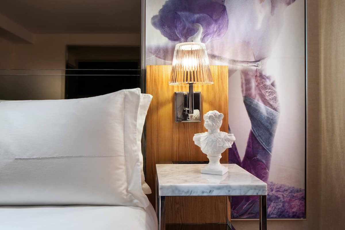 A hotel room with a bed, a marble nightstand, a wall-mounted lamp, and a large floral artwork.