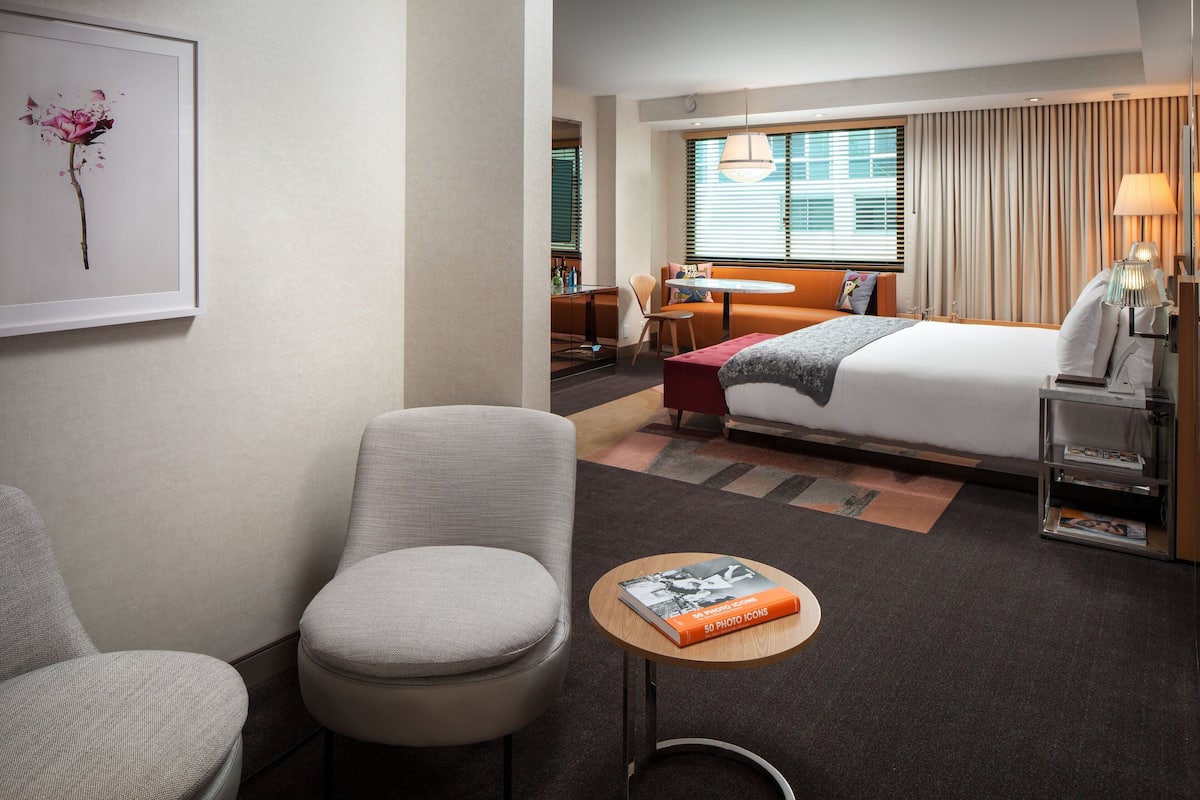 A hotel room with a bed, a chair, a small table with a book, a lamp, and a window with blinds.