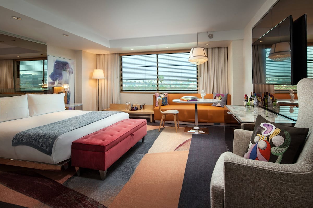 A modern hotel room with a large bed, a red ottoman, a sofa, a dining table, and a TV.