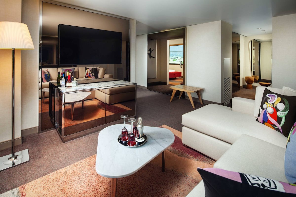 A modern hotel room with a flat-screen TV, a sofa, a coffee table with decorative items, and a view of the city.