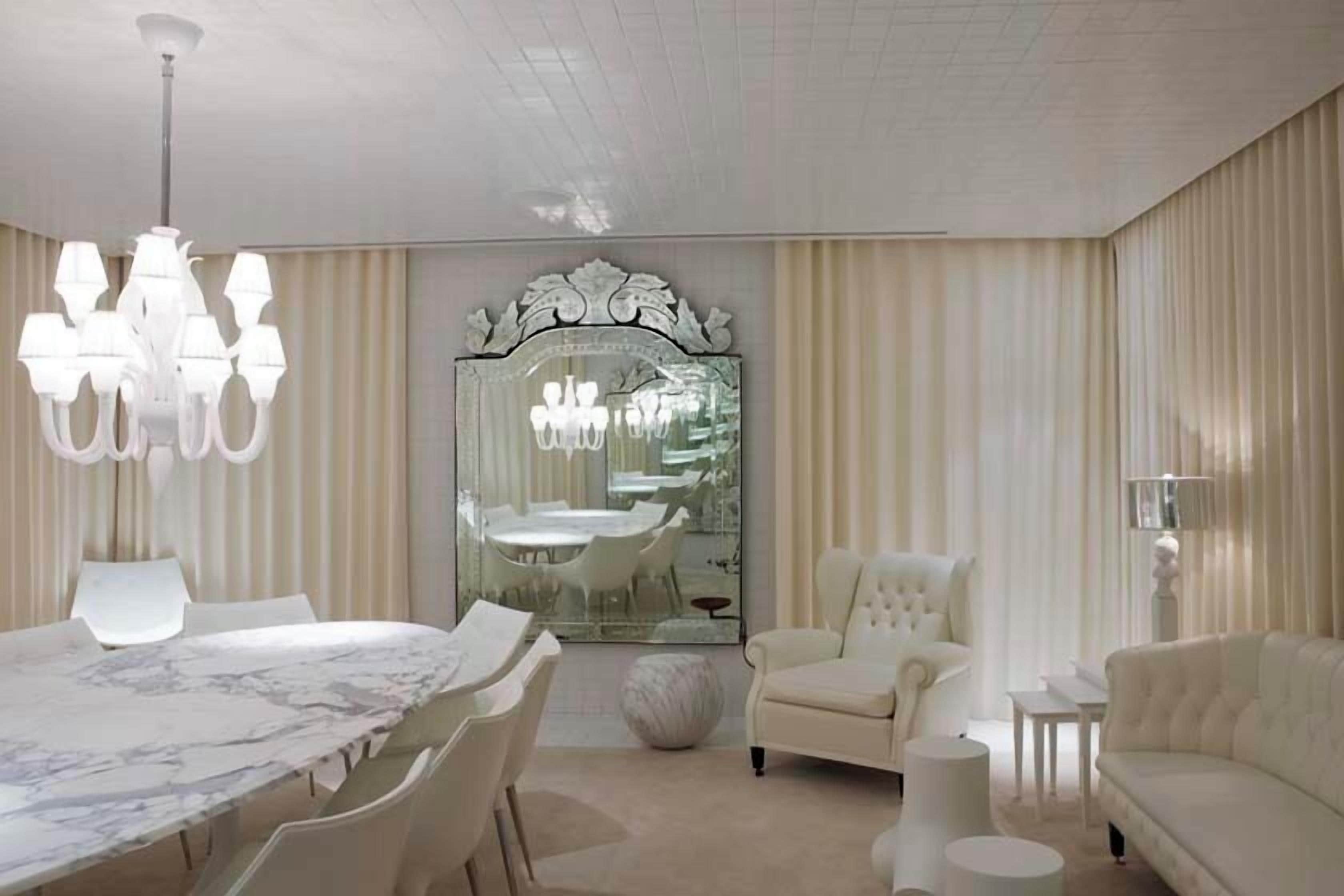 A modern living room with a marble dining table, white armchairs, a sofa, a chandelier, and a large mirror with an ornate frame.