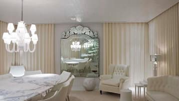 A modern living room with a marble dining table, white armchairs, a sofa, a chandelier, and a large mirror with an ornate frame.