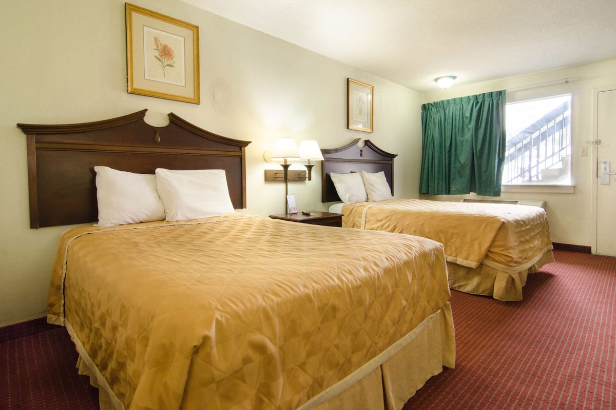 A hotel room with two beds, a nightstand, a lamp, a window with curtains, and framed pictures on the wall.