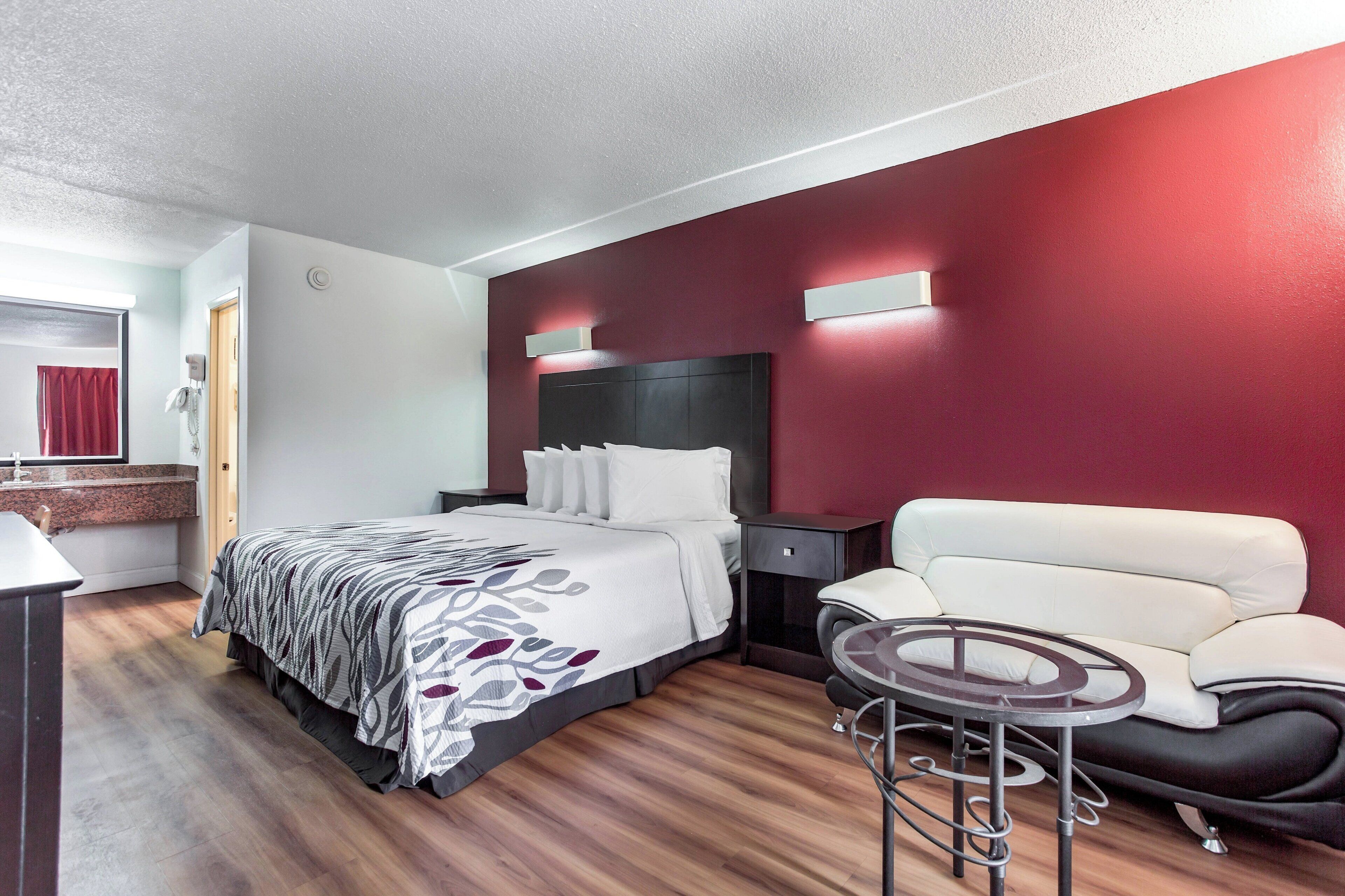 Una habitación de hotel con cama, sofá, una mesita, espejo y una pared de acento en rojo.