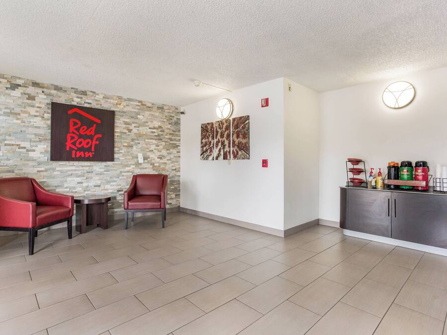 Een Red Roof Inn lobby met stenen muur, twee rode stoelen, een kleine tafel, kunstwerken en een kitchenette met koffiezetapparaten.