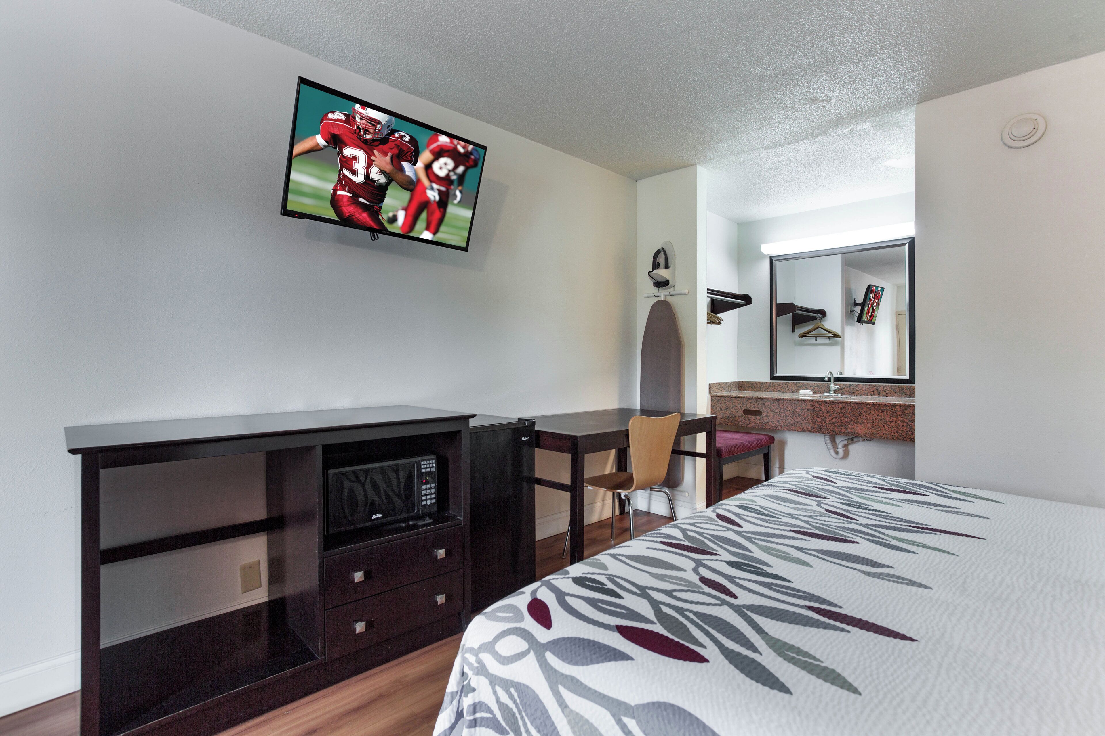 Habitación de hotel con cama, televisión montada en la pared, escritorio con silla, cómoda y espejo sobre la cómoda.