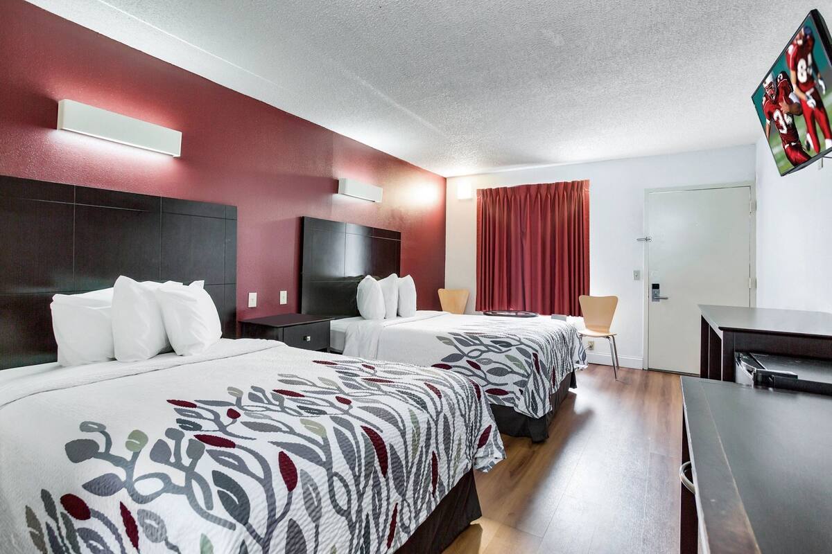 A hotel room with two beds, a desk, a chair, a TV, and a door.