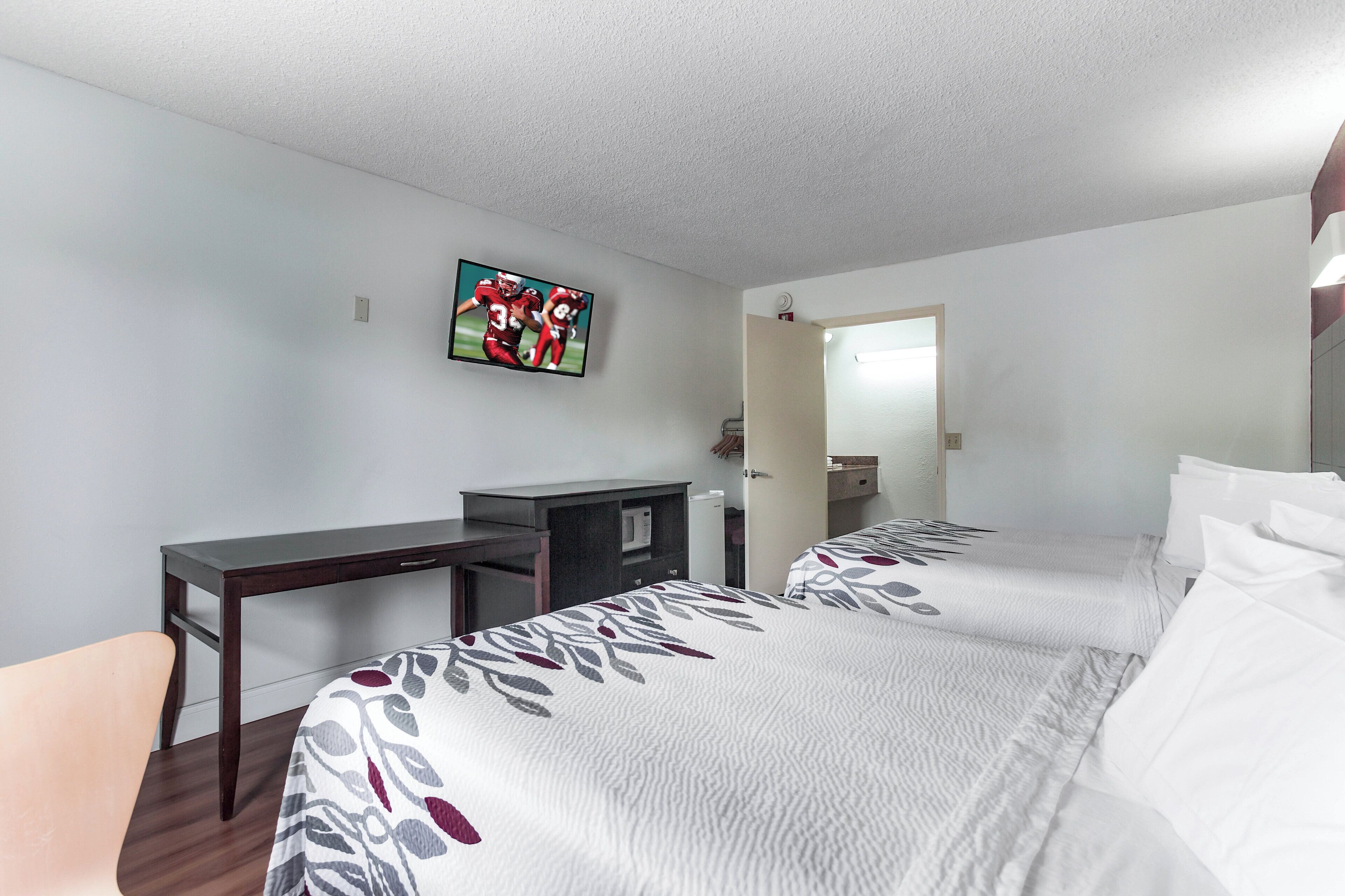 A hotel room with a bed, a desk, a television mounted on the wall, and a door leading to a bathroom.