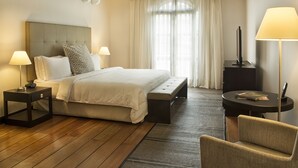 A hotel room with a large bed, two bedside tables with lamps, a bench, a chair, a small table with a lamp, and a large window with curtains. - Movich Casa del Alferez (Cali)