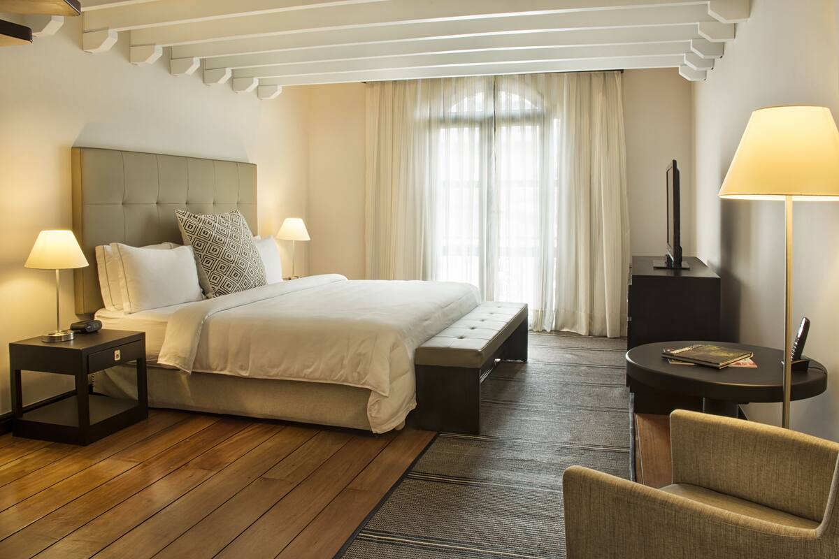 A hotel room with a large bed, two bedside tables with lamps, a bench, a chair, a small table with a lamp, and a large window with curtains.