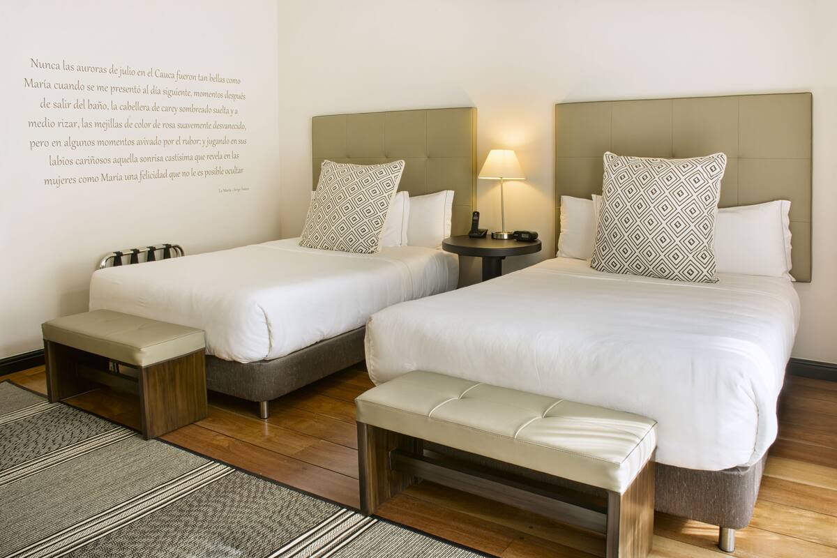 A hotel room with two beds, a bedside table with a lamp, a bench, and a patterned wall decoration.