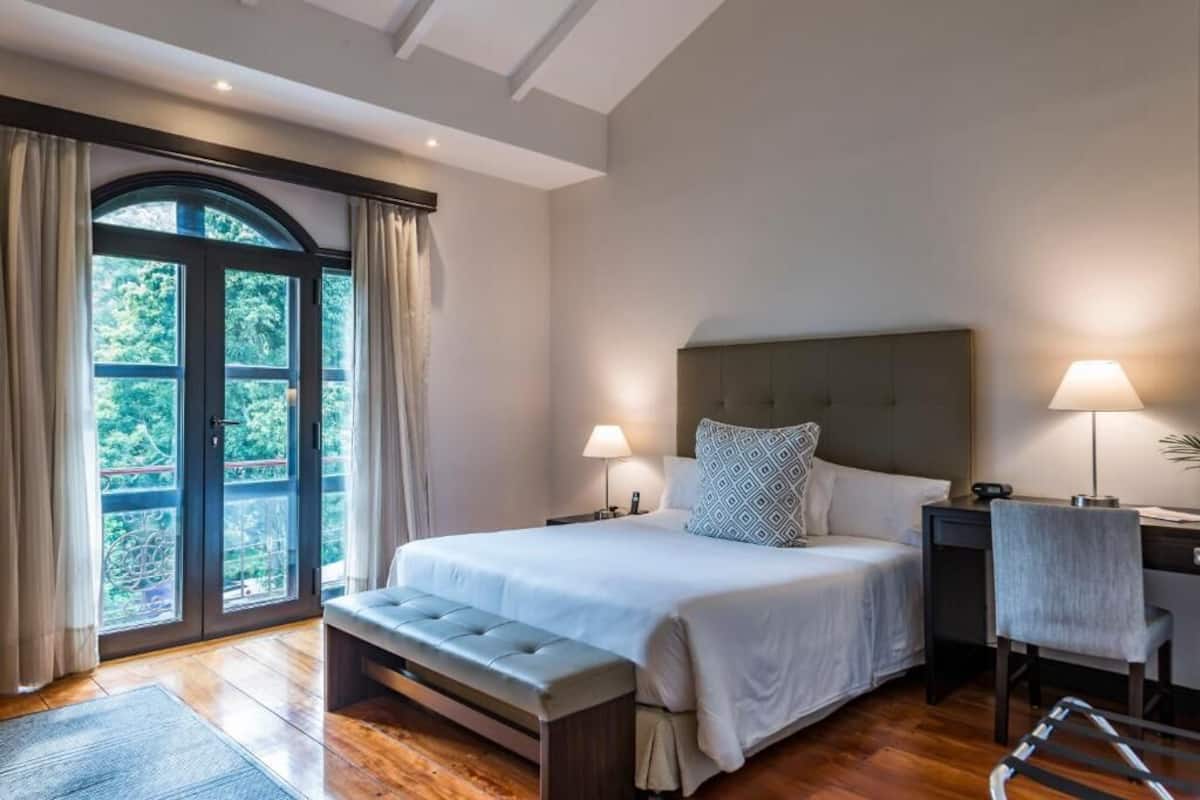A bedroom with a large bed, two bedside tables with lamps, a bench at the foot of the bed, and a view through a glass door.