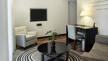 A modern hotel room with a round glass coffee table, a sofa, a chair, a desk with a lamp, a TV, and a potted plant.