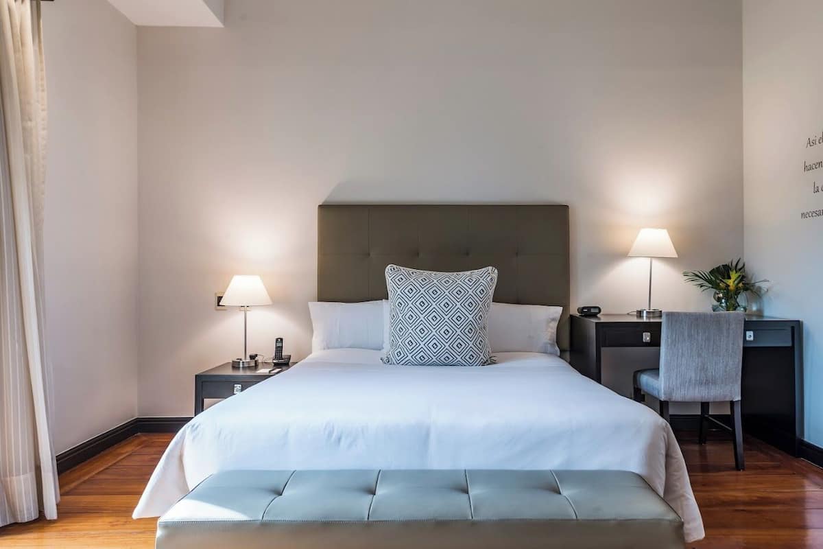 A hotel room with a large bed, two bedside tables, a desk with a chair, and a window with curtains.