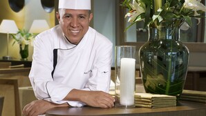 A chef in a white uniform seated at a table with a vase of flowers and a candle. - Movich Casa del Alferez (Cali)