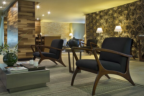 A hotel lobby with two armchairs, a coffee table, a vase with flowers, a fireplace, and patterned wallpaper.