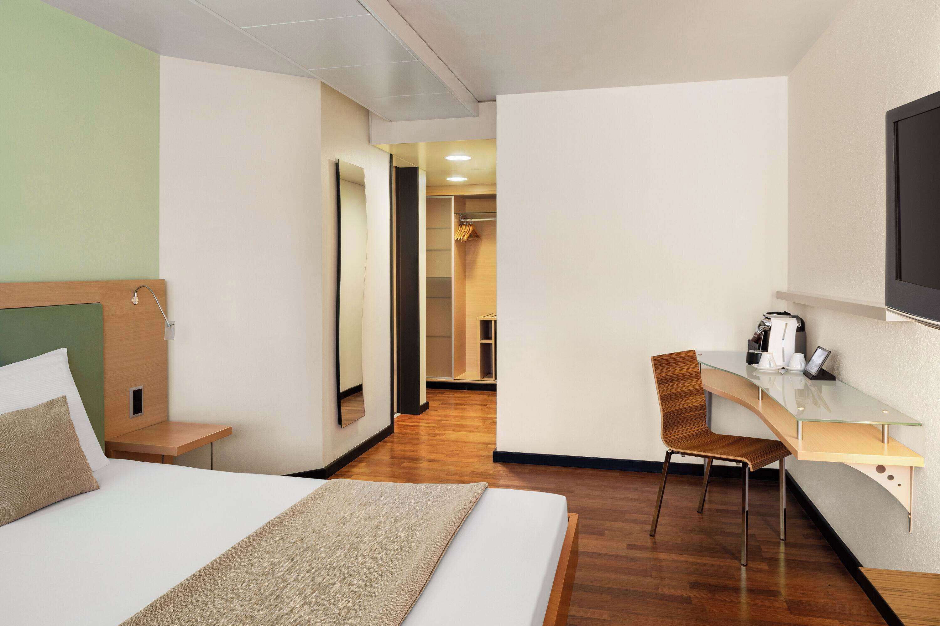 Ein Hotelzimmer mit Bett, Schreibtisch, Stuhl, Spiegel und Fernseher.