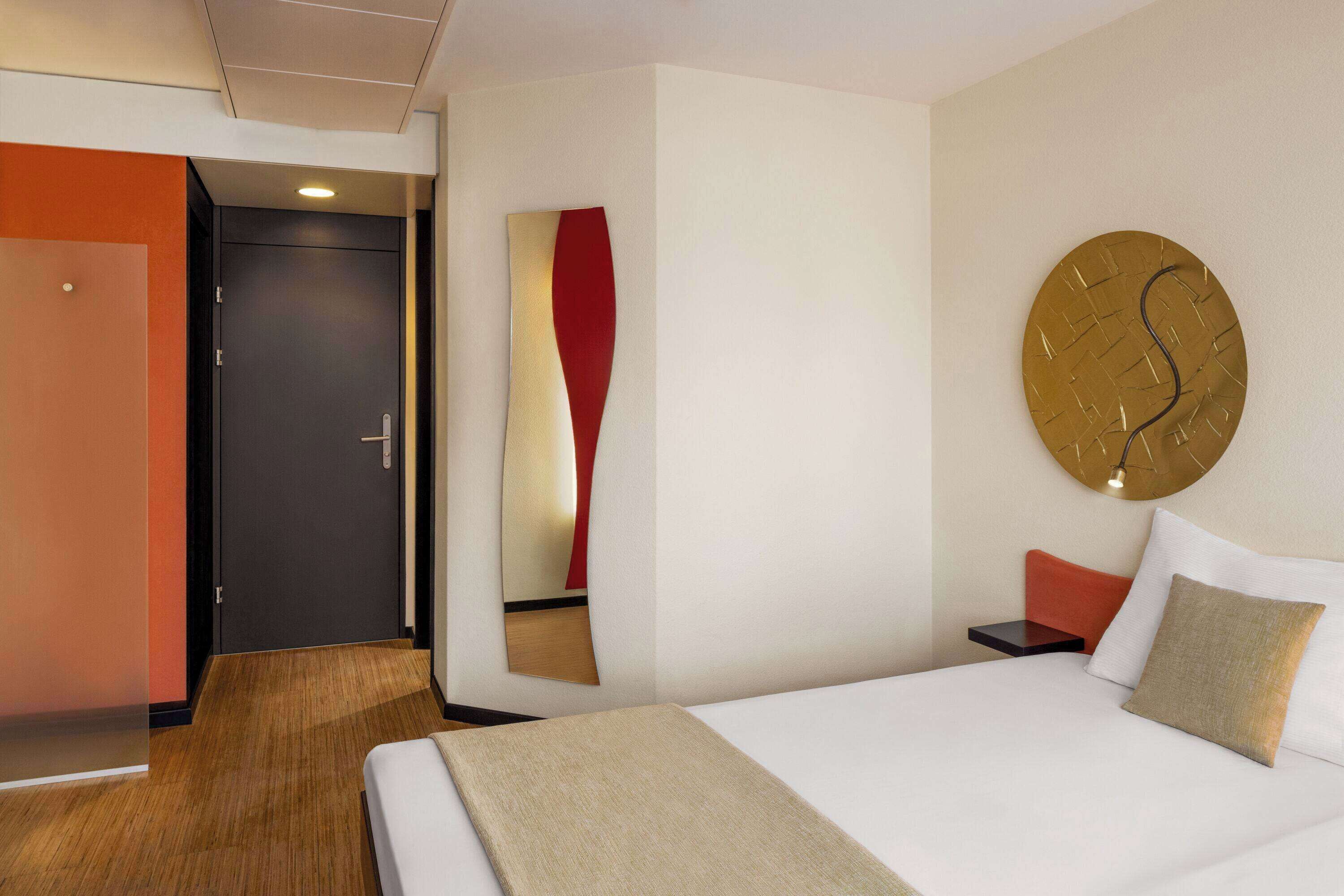 A hotel room with a bed, a bedside table, a wall clock, and a door.