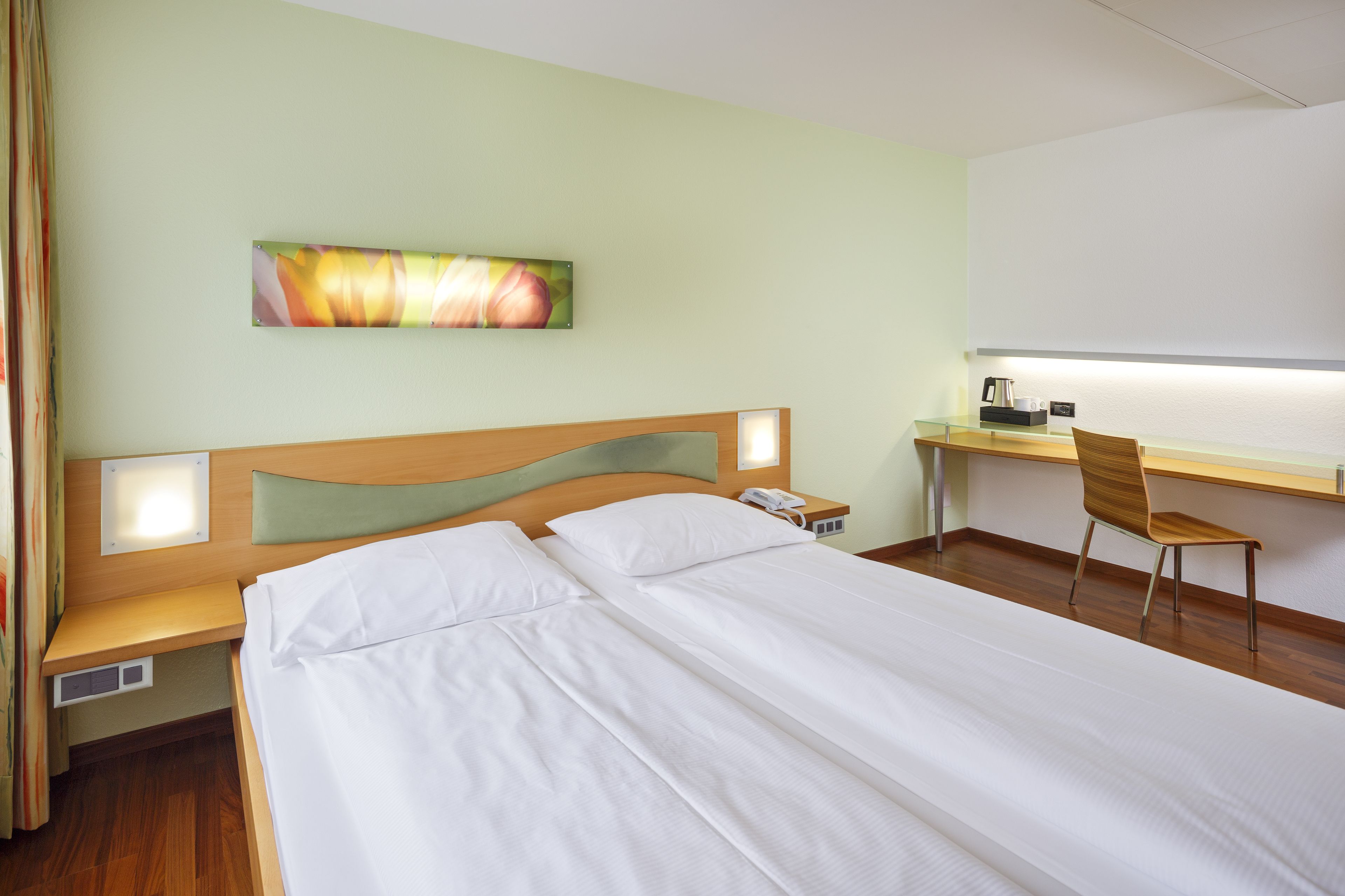 Habitación de hotel con cama, escritorio con silla, mesita de noche y un cuadro colgado en la pared.