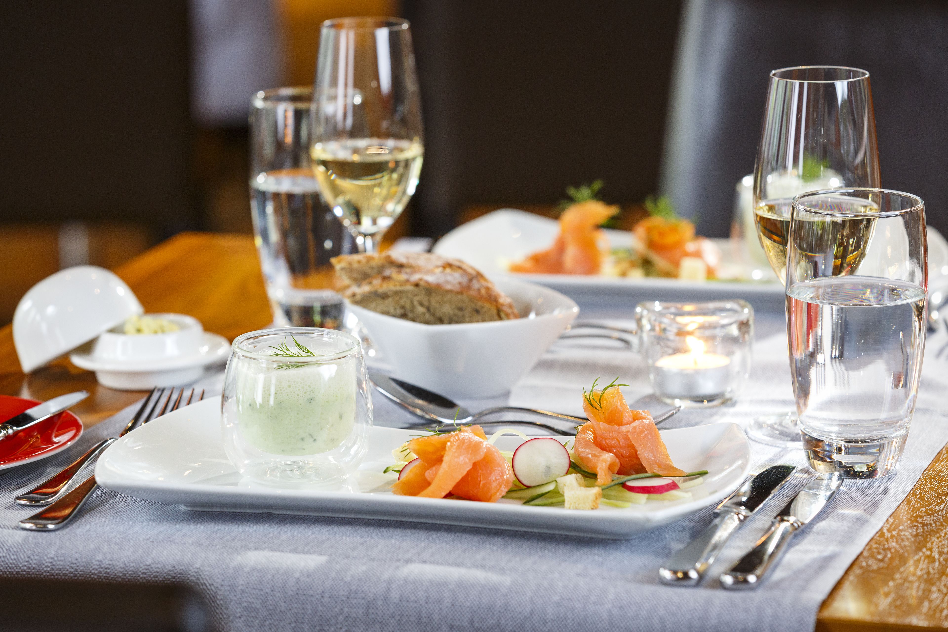 Una mesa de comedor dispuesta con platos de comida, copas de vino blanco y una vela.