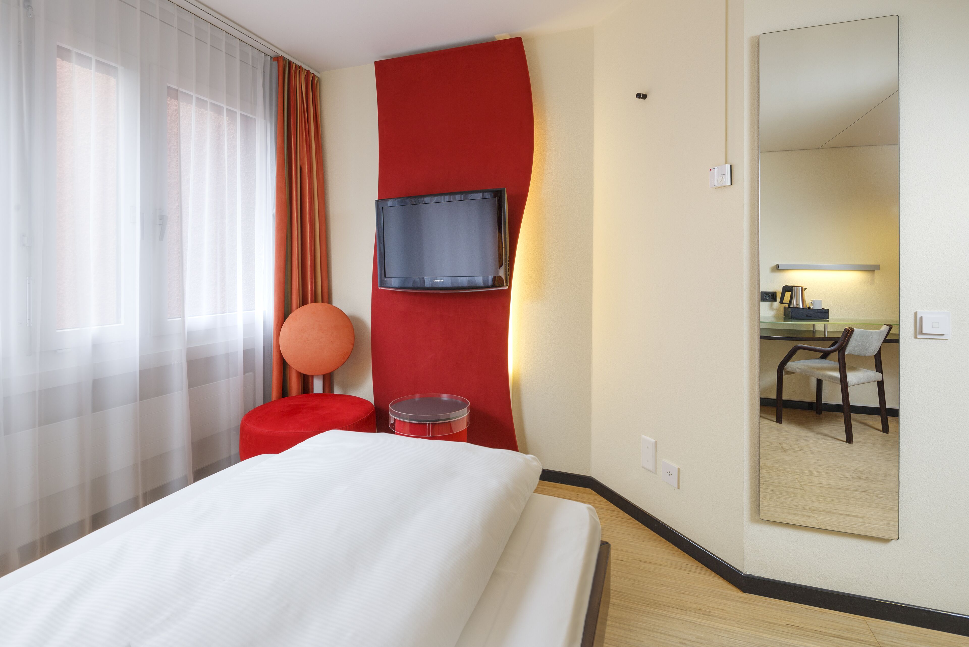 Habitación de hotel con cama, pared decorada en rojo, televisor montado, silla roja redondeada, una mesita y un espejo que refleja un escritorio y una silla.