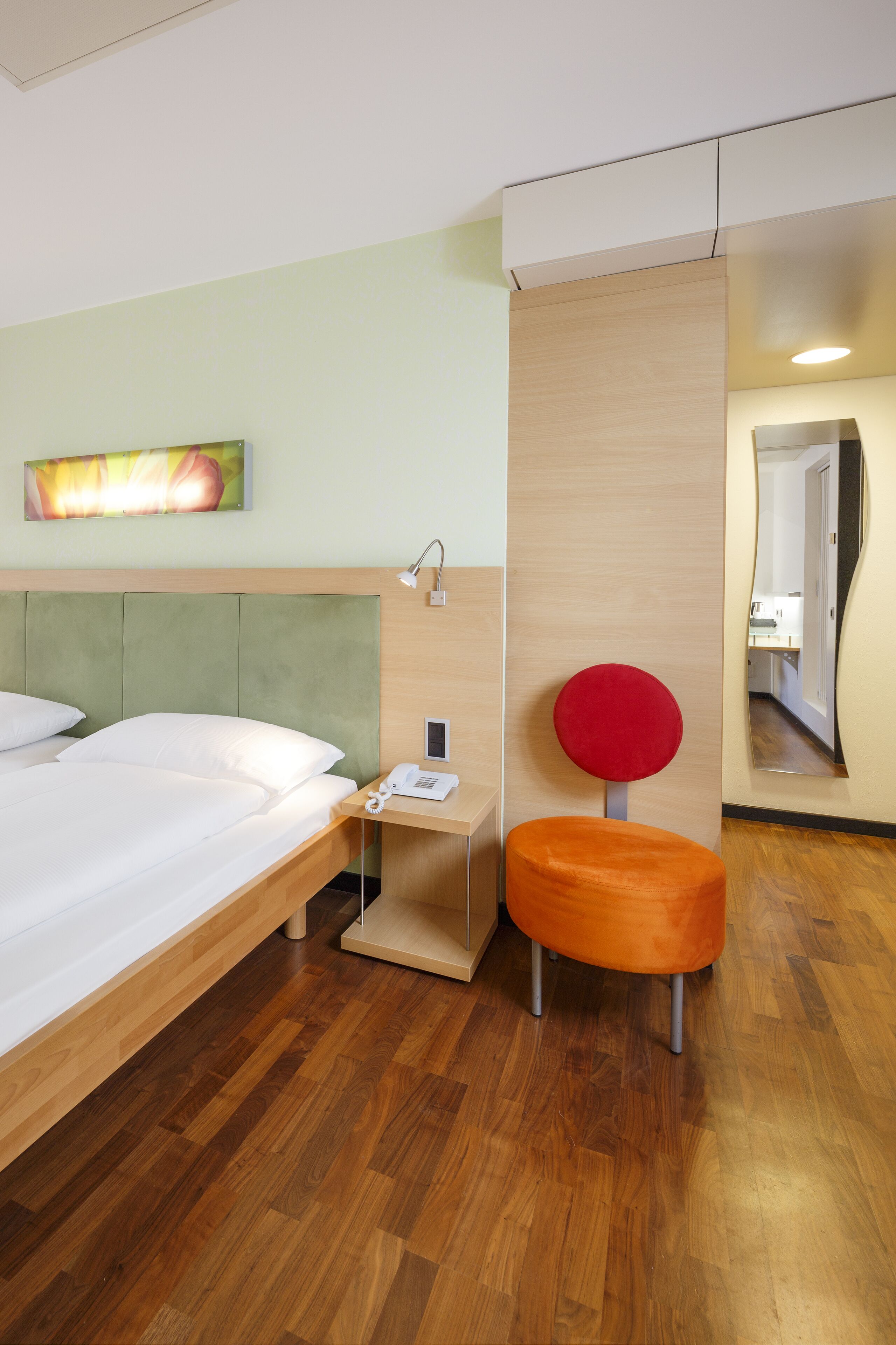 A hotel room with a bed, a nightstand, a red chair, a mirror, and a painting on the wall.