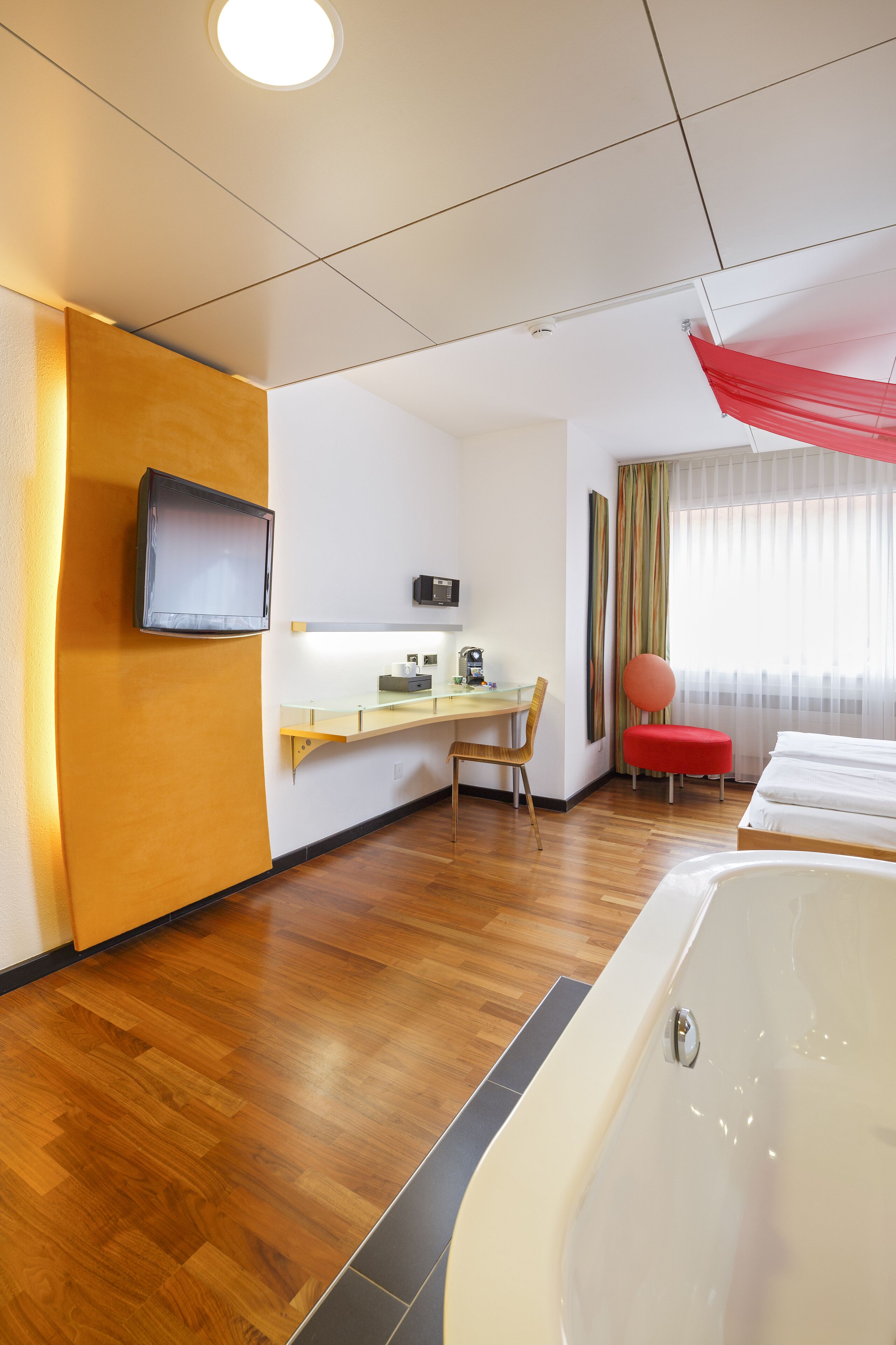 Een hotelkamer met een houten vloer, een bed, een bureau met stoel, een aan de muur bevestigde televisie en een raam met gordijnen.