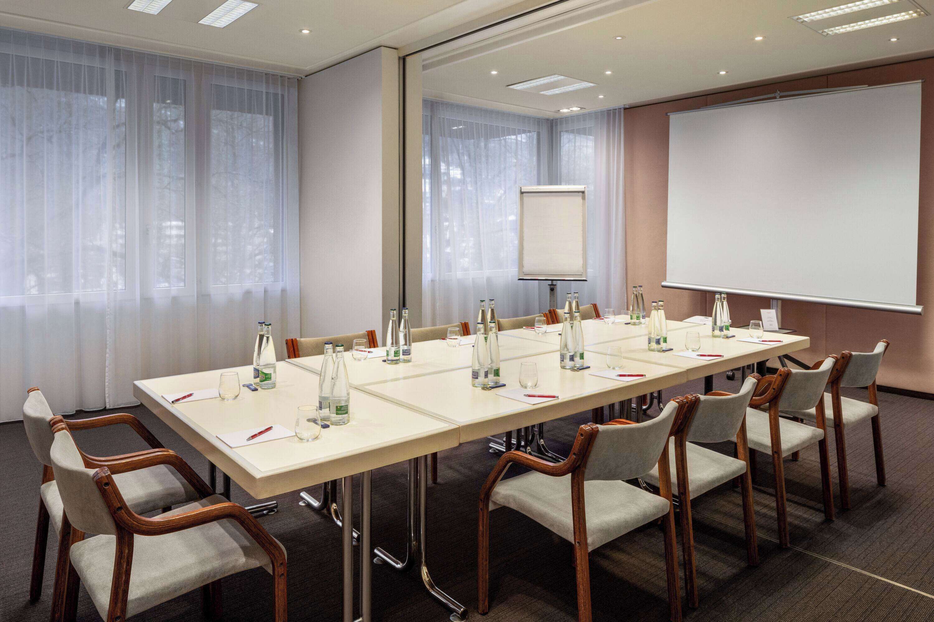 A conference room with a long table, chairs, a projector screen, and a large window with sheer curtains.