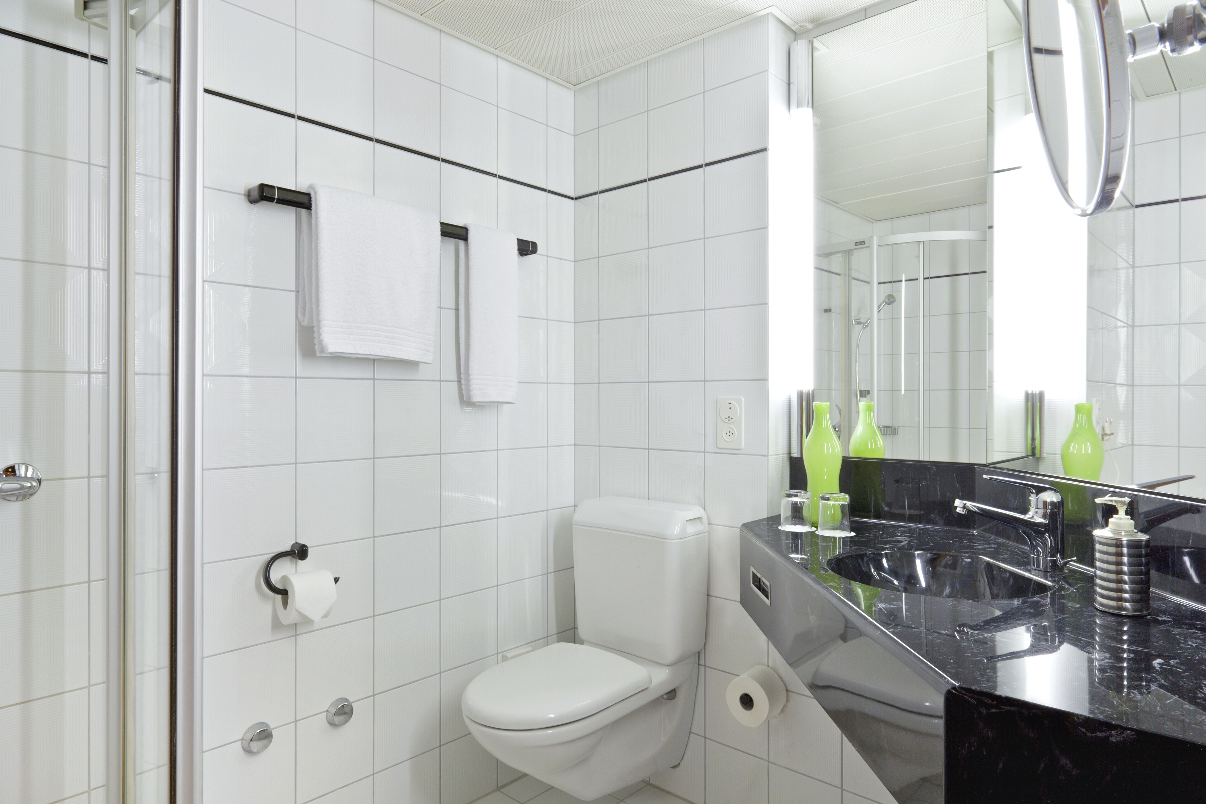 Uma casa de banho moderna com azulejos brancos, sanita, lavatório com bancada preta e espelho.