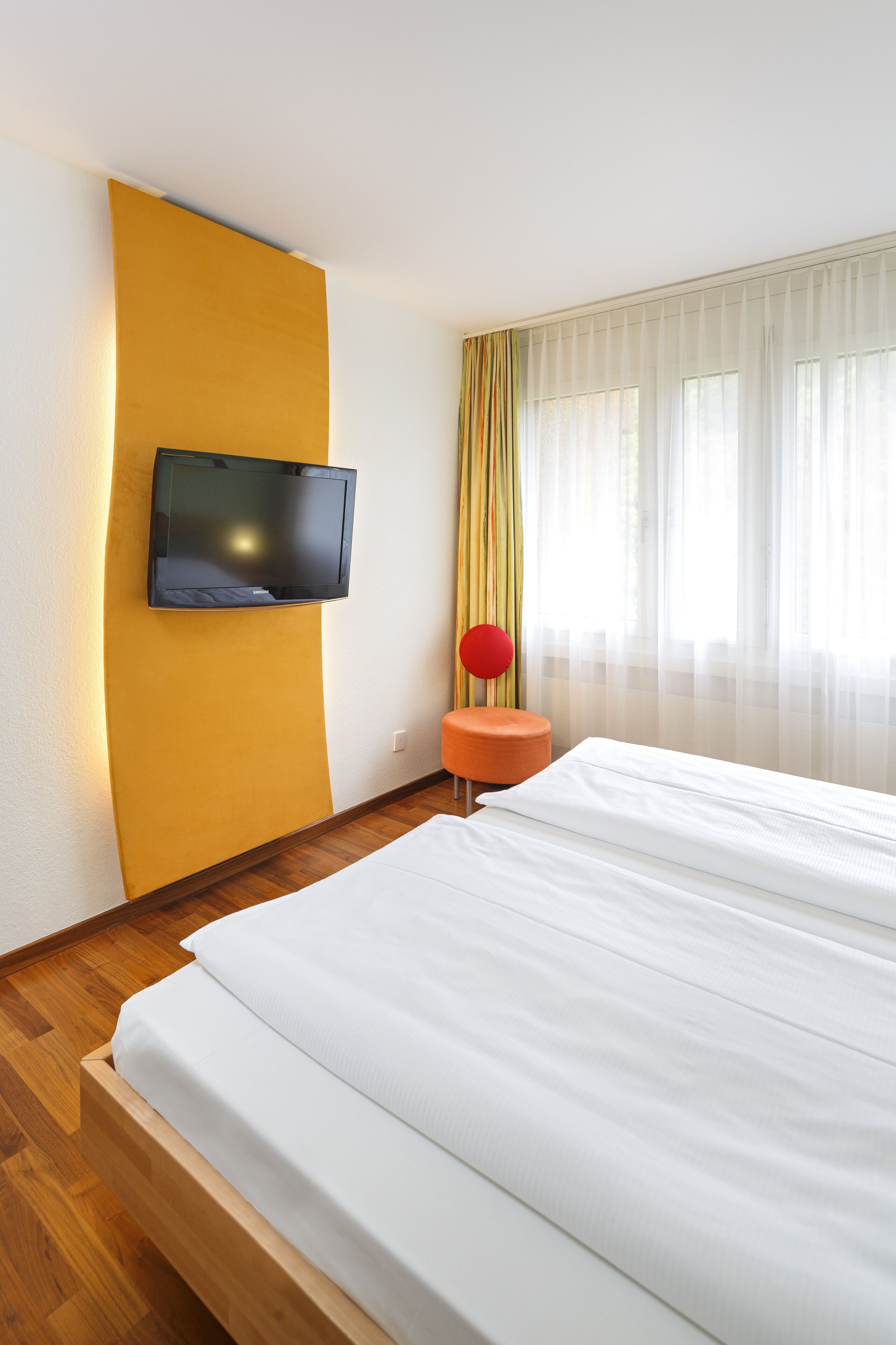Une chambre d’hôtel avec un lit, un téléviseur fixé au mur, un petit tabouret orange et une fenêtre avec des rideaux légers.