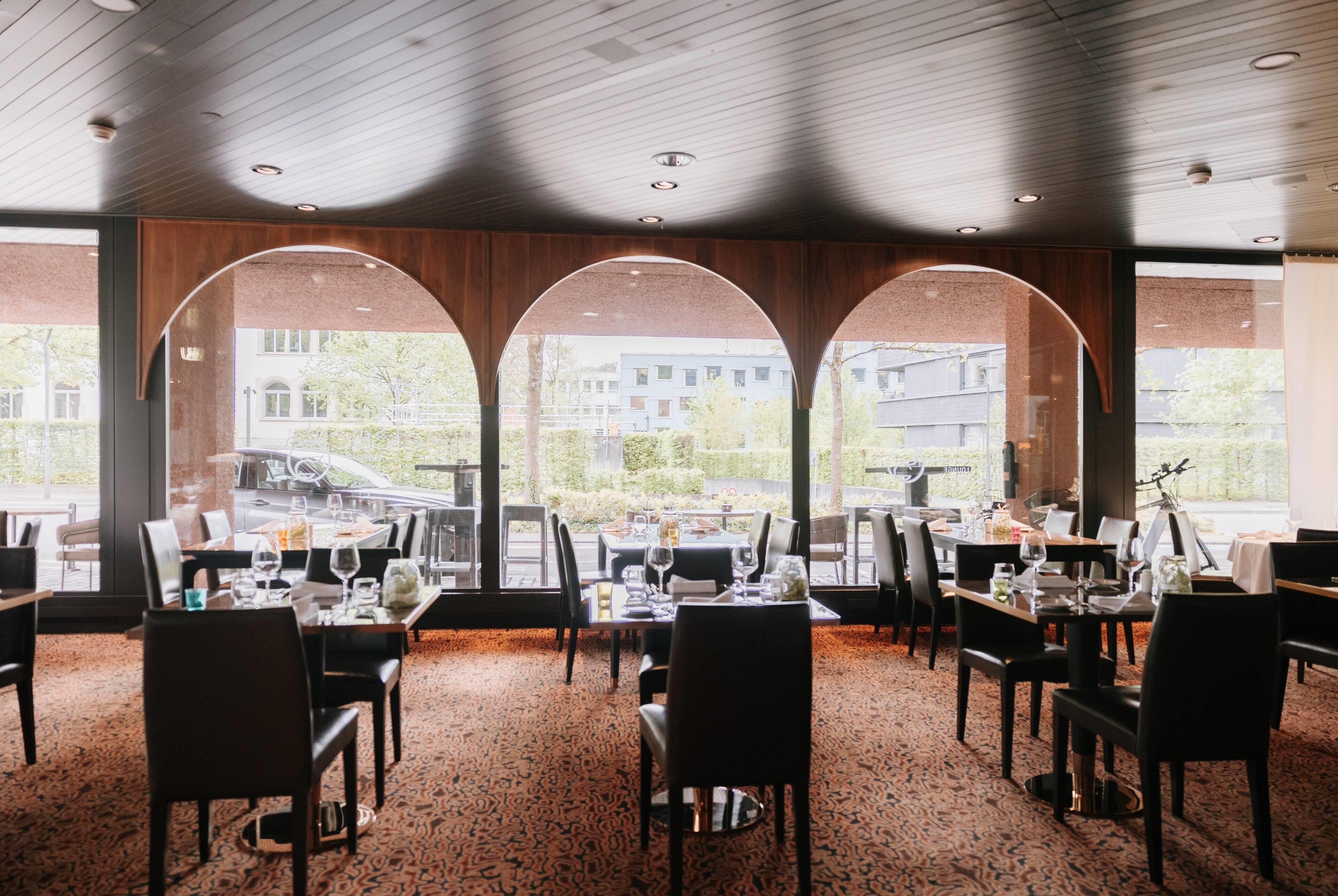 A restaurant with tables set for dining, large windows with arched frames, and a view of buildings outside.