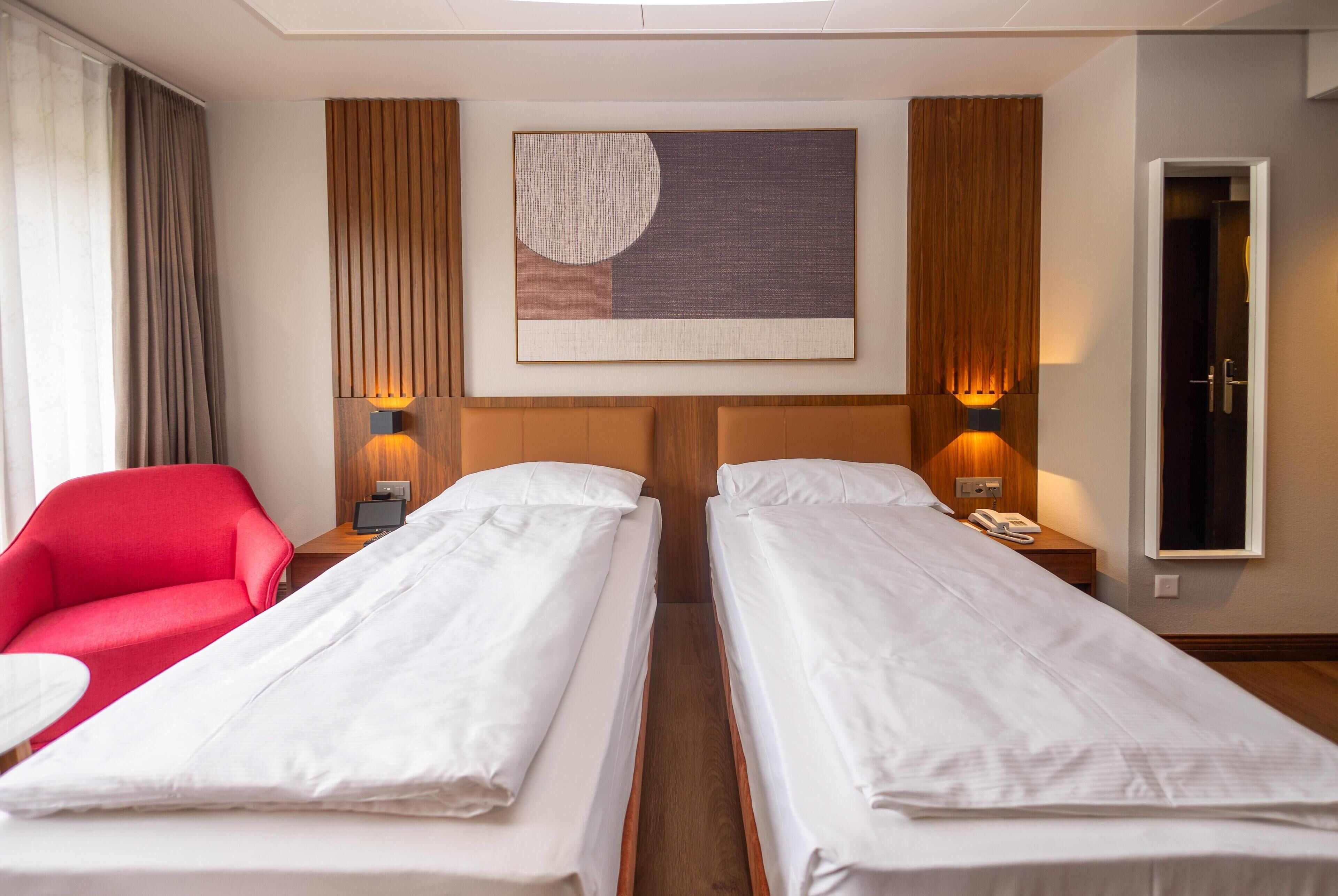 A hotel room with two single beds, a red chair, a wooden headboard, a wall-mounted painting, and bedside lamps.