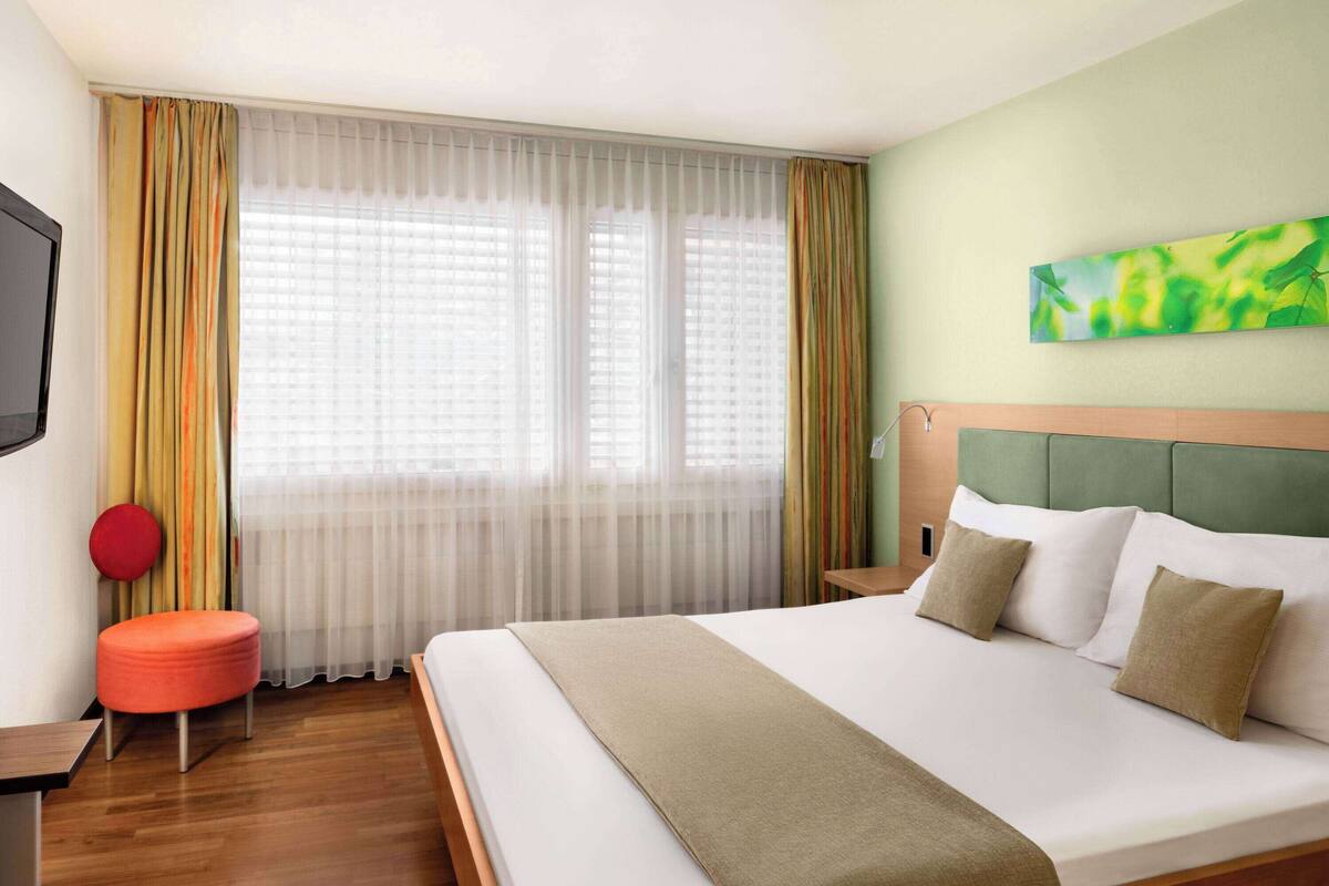 Een hotelkamer met een groot bed, een rode poef, een televisie en een schilderij aan de muur.