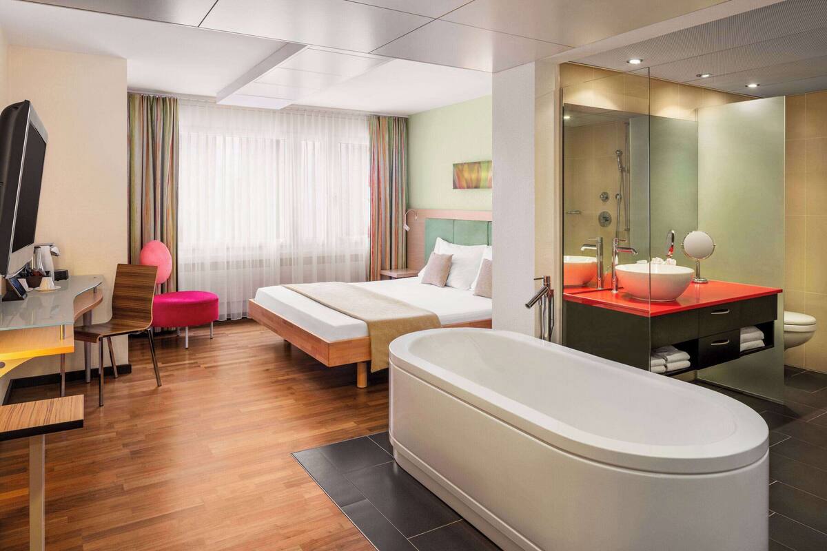 Een moderne hotelkamer met een groot bed, een apart zithoekje met een stoel, een bureau, een ruim bad en een badkamer met spiegel en wastafel.