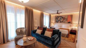 A hotel room with a blue sofa, a patterned armchair, a wooden coffee table, a bed with white bedding, and a painting on the wall.