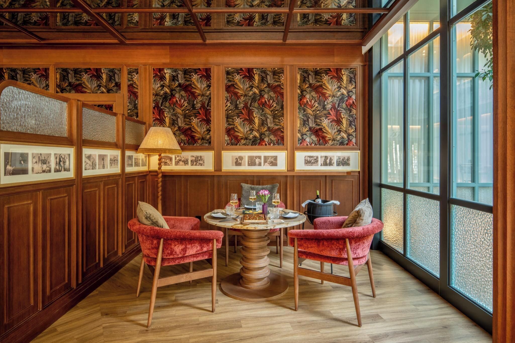 A room with a wooden ceiling, a large window, a round table with chairs, and framed pictures on the walls.