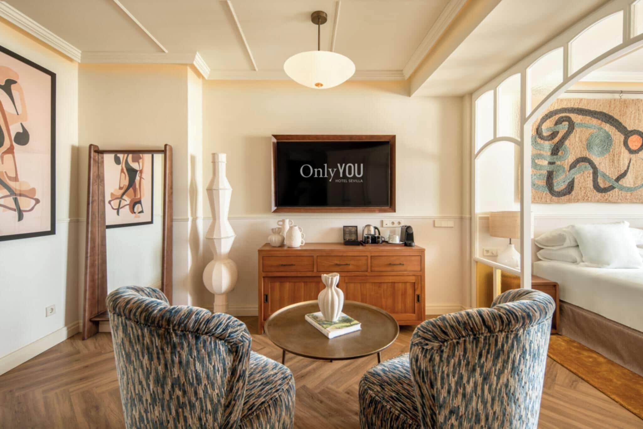 Habitación de hotel con televisión de pantalla plana, sofá, mesa de centro y cama con cabecero decorativo.