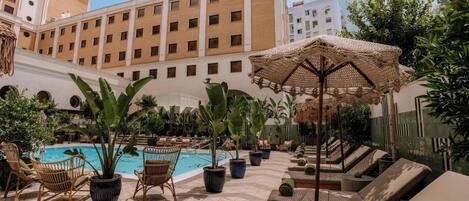 Zona de piscina de hotel con sillones de descanso, plantas en macetas y un edificio grande al fondo.