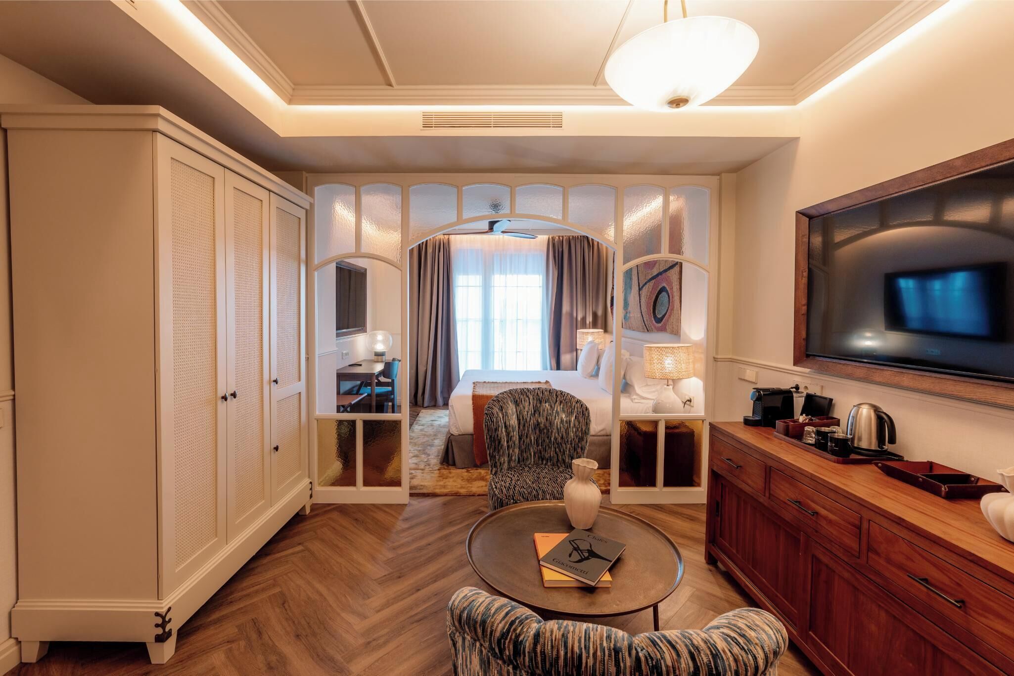 A hotel room with a bed, a seating area with a table, a TV, and a wardrobe.