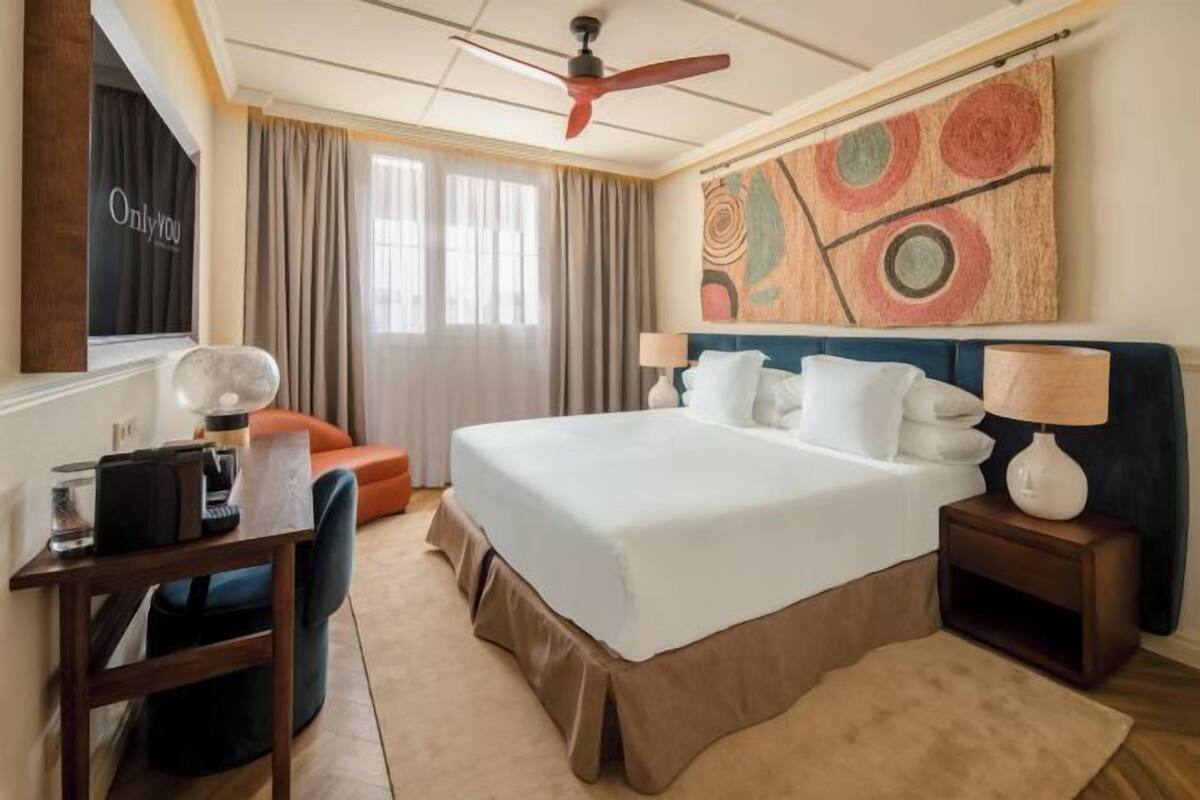 Habitación de hotel con una cama grande, un escritorio con una silla, una mesita de noche con una lámpara, un ventilador de techo y una ventana con cortinas.